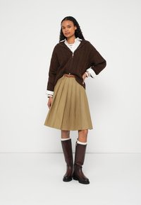 Hnedý sveter s pleteným vzorom, na zips, krémová košeľa s golierom, plisovaná khaki sukňa a hnedé čižmy po kolená s bielymi detailmi.