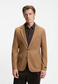 Blazer in lana marrone con due tasche frontali, rever a rotondo e chiusura con bottone. Indossato sopra una polo scura, con una texture sottile.