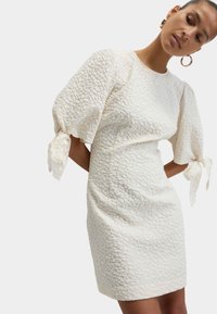 Bílá texturovaná šaty s nafouknutými rukávy a stužkami na zápěstí. Přiléhavý silueta s jemným vzorem po celém materiálu. Krátká délka.