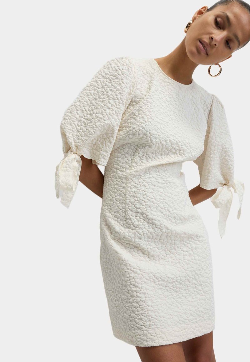 Bílá texturovaná �šaty s nafouknutými rukávy a stužkami na zápěstí. Přiléhavý silueta s jemným vzorem po celém materiálu. Krátká délka.
