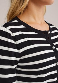 Cardigan rayé noir et blanc avec un col rond, des manches bouffantes et des accents de boutons en perles, présentant une texture douce et tricotée.