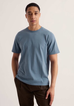 Homme debout devant un fond clair, portant un t-shirt bleu à manches courtes et un pantalon marron foncé, regardant directement la caméra.