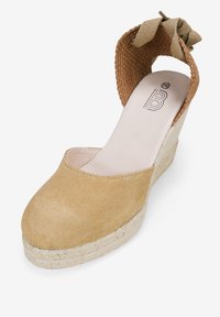 Zapato de tacón cuña de ante beige con una parte superior de tejido texturizado y un detalle de tela atado en la parte posterior, que presenta una plantilla de yute trenzado.
