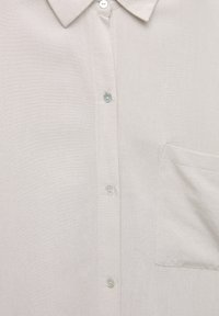 Chemise beige clair à boutons avec col et poche poitrine, présentant quatre boutons visibles sur la patte de boutonnage avant.