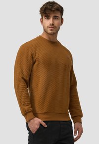 Brauner, strukturierter Pullover mit rundem Ausschnitt und gerippten Bündchen. Ausgestattet mit einem kleinen Logodetail auf der Brust, kombiniert mit schwarzen Hosen.