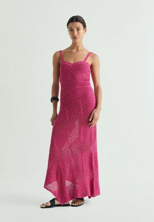 Mujer con un vestido maxi de crochet sin mangas de color rosa brillante, sandalias negras y una pulsera negra, de pie frente a un fondo claro y liso.