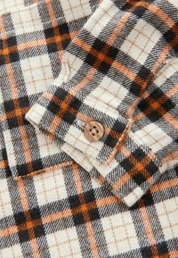 Flanelinė marškiniai su dryžuotu raštu juodos, baltos ir oranžinės spalvų, su vienu rudu mygtuku ir minkšta tekstūra.