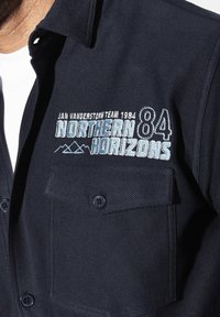 Dunkelblaues Hemd mit Knöpfen aus strukturiertem Stoff, das ein gesticktes Design mit "Northern Horizons 84" und einem Bergmotiv auf der Brusttasche zeigt.