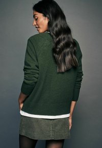 Mørkegrøn strikket sweater med ribbede manchetter og detalje på ryggen, lagdelt over en hvid skjorte og olivengrøn velour-nederdel, mod en neutral baggrund.