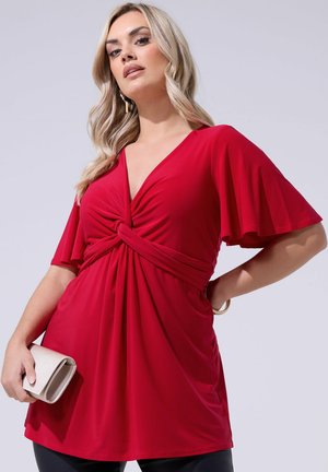 Woman with long blonde hair wearing a red twist-front tunic with flutter sleeves, holding a small beige clutch against a neutral background.