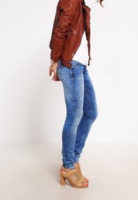 Chaqueta de cuero marrón, camisa blanca, jeans skinny azules y sandalias de tacón alto color tostado con un diseño perforado.