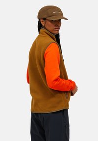 Braune Fleece-Weste mit orangefarbenen langen Ärmeln, strukturiertem Stoff, hohem Kragen und Seiten Taschen. Dunkle Hose und braune Mütze mit Logodetails.