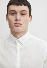 Camisa blanca de lino con cuello clásico, que presenta botones claros y una textura sutil. Pequeño logotipo bordado en el lado izquierdo.