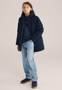 Tengerészkék kapucnis parka, elöl cipzárral, tengerészkék pulóverrel és világoskék farmernadrággal párosítva. Az öltözethez barna talpú sportcipők tartoznak.