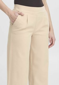 Pantaloni beige a gamba larga con una texture liscia, dotati di una vita elastica alta e tasche laterali con pieghe sottili.