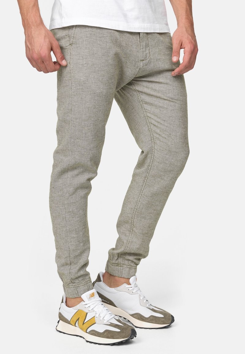 Homme portant un pantalon de jogging gris clair et des baskets blanches avec des accents jaunes et gris, debout devant un fond uni.