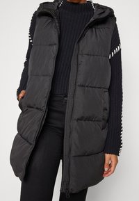 Czarna pikowana kamizelka z kapturem, zapinana na zamek z przodu, z bocznymi kieszeniami i pikowanym wzorem. Nosi się ją na granatowym swetrze dzianym.