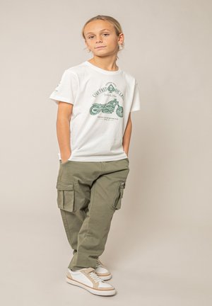 Enfant les mains dans les poches portant un t-shirt blanc à motif moto, un pantalon cargo vert olive et des baskets blanches avec des accents beige.