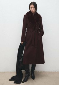 Bordeauxrode wollen jas met een kunstbontkraag, gëceintuurd in de taille; gedragen met zwarte kniehoge laarzen en een zwarte sjaal losjes over de schouder.