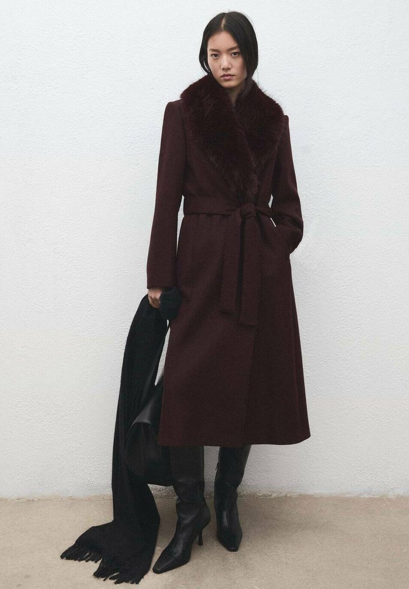 Bordeauxrode wollen jas met een kunstbontkraag, gëceintuurd in de taille; gedragen met zwarte kniehoge laarzen en een zwarte sjaal losjes over de schouder.