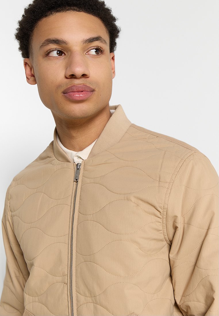 Giacca bomber trapuntata beige con zip frontale, colletto a costine e un sottile motivo a onde, indossata sopra una camicia con colletto.