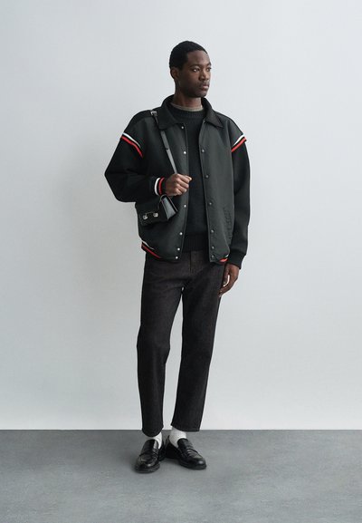 Must mustrepp, millel on punased ja valged ribaaktsendid. Tasane kampsun, tumedad teksad, valged sokid ja mustad loaferid. Must ristseljakott.