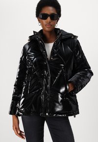 Black glossy puffer jacket with a hood, diamond quilting, and gold hardware accents. Model wearing it with hands in pockets, paired with sunglasses.