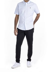 Camisa de botones de manga corta en azul claro, pantalones negros y zapatillas blancas. La camisa tiene un pequeño logo en el pecho. Textura suave.