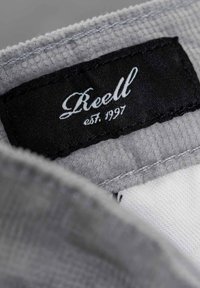 Gray fabric tag with black text reading "Reell" and "est. 1997." Background shows soft gray textured material and white fabric.