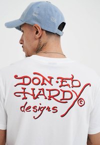Bílý tričko s krátkým rukávem, na zadní straně s vyšitým textem "DON ED HARDY designs" v červené a černé barvě; bavlněný materiál; kulatý výstřih.