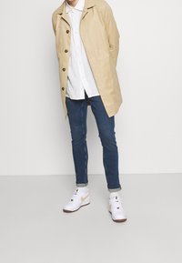 Trench coat ligero beige con botones oscuros, camisa blanca con cuello, jeans azul slim-fit y zapatillas blancas con acentos beiges.