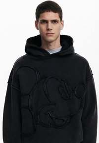 Sort hættetrøje med et fremtrædende monochromt broderet Mickey Mouse-design. Fremstillet af blødt stof med en afslappet pasform og stor hætte.