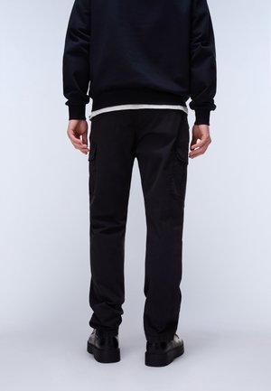 Persona de espaldas, vestida con pantalones cargo negros, sudadera negra, camiseta interior blanca y zapatos negros sobre un fondo liso.