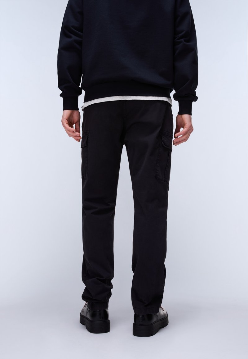 Personne debout de dos portant un pantalon cargo noir, un sweat-shirt noir, un maillot blanc en dessous et des chaussures noires sur un fond uni.
