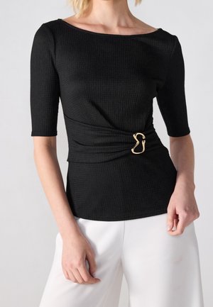 Woman wearing a black textured top with elbow-length sleeves and a gold decorative buckle detail, paired with white pants.