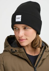 Čierna rebrovaná čiapka s bielou obdĺžnikovou nálepkou s logom "NORTH BEND". Nosí sa s hnedou bundou na zips, odhaľujúcou textúrovanú tkaninu.