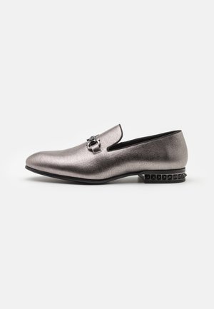 Mocassins argentés métalliques avec une finition texturée, présentant une boucle décorative et un talon noir clouté, mettant en avant un design élégant et épuré.