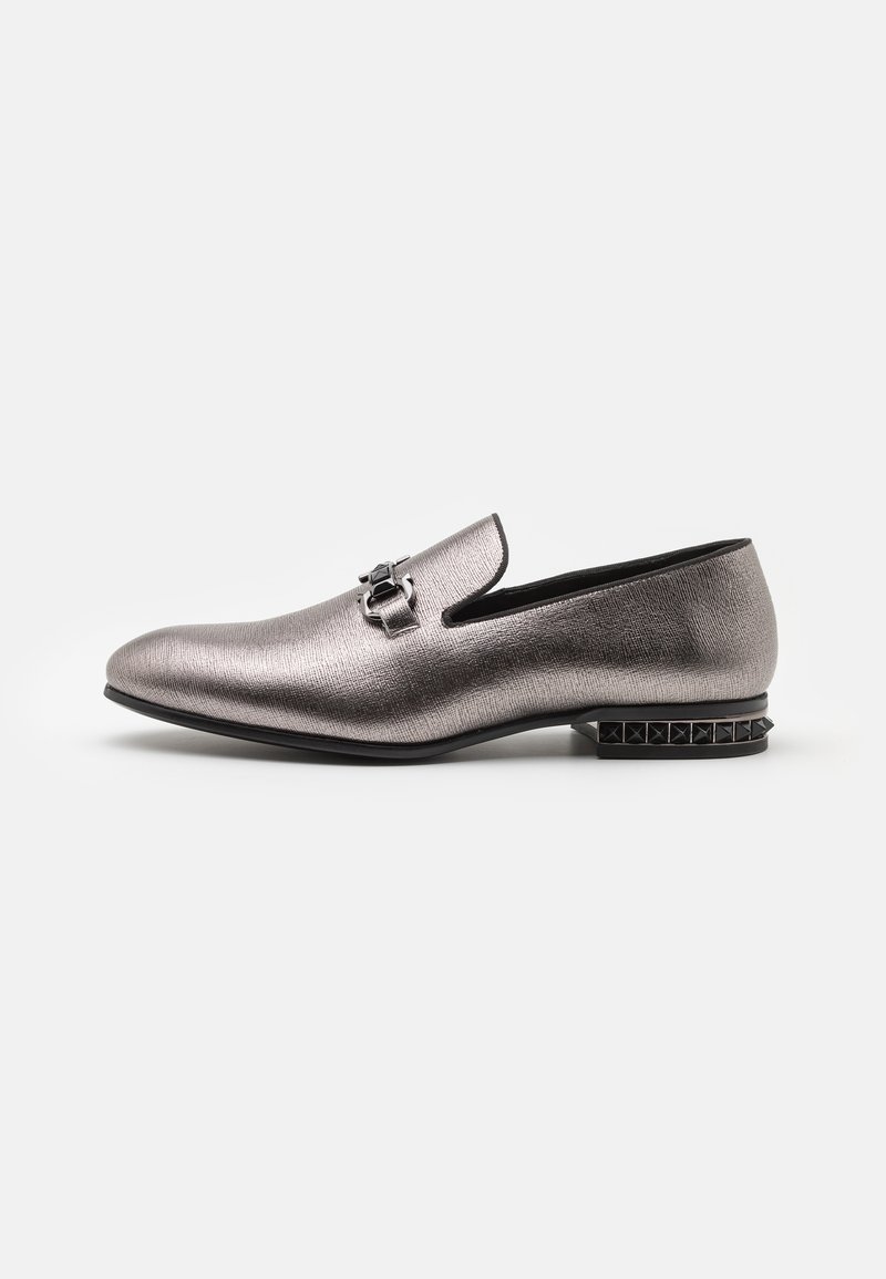 Mocassins argentés métalliques avec une finition texturée, présentant une boucle décorative et un talon noir clouté, mettant en avant un design élégant et épuré.