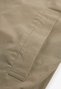 Tessuto beige con una texture liscia, caratterizzato da una tasca laterale con una pattina dritta e linee di cucitura visibili. Design semplice e funzionale.