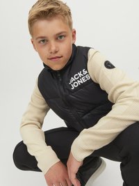 Črn napihnjen suknjič z belim logotipom "JACK & JONES", bež rebraste dolge rokave, črne hlače, priklonjen položaj, nevtralna ozadje.