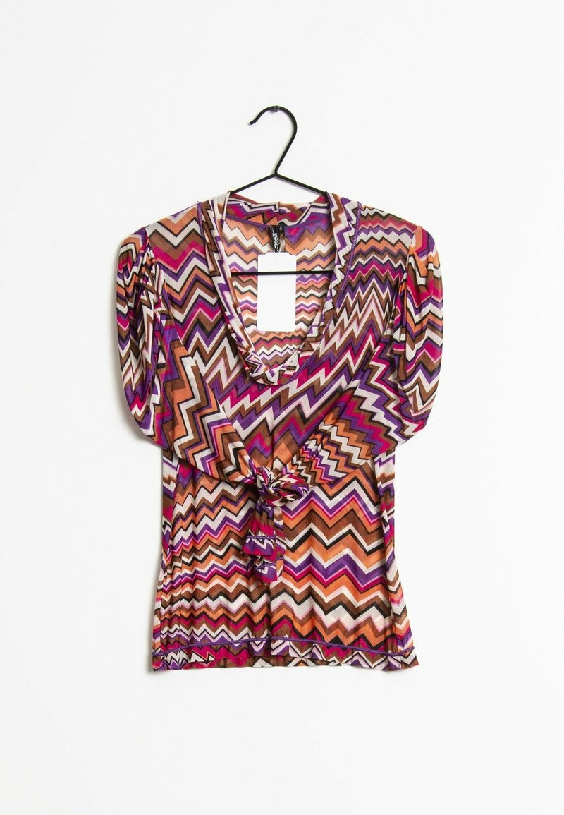 Blouse à manches longues avec motif en zigzag dans des tons violet, orange et marron, nouée à la taille, suspendue sur un cintre noir.