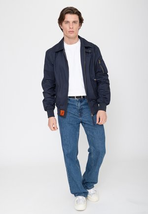 Young man standing, wearing navy bomber jacket, white t-shirt, blue jeans, black belt, and white sneakers against plain white background.