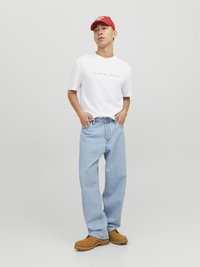 Weißes T-Shirt mit schwarzem Text, hellblaue weite Jeans und braune Arbeitsstiefel, ergänzt durch eine rote Baseballkappe. Einfarbiger Hintergrund.