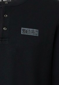 Zwarte lange mouwen henley shirt gemaakt van ribstof, met drie zwarte knopen en een grijs label met "s.Oliver Elevated Casual Est. 1969."