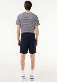 Homme debout de dos portant un t-shirt rayé noir et blanc, un short bleu marine, des chaussettes blanches et des chaussures blanches sur un fond blanc.