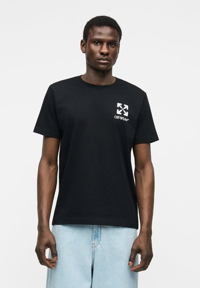 Νέος άνδρας στέκεται φορώντας μαύρο t-shirt Off-White με λευκό λογότυπο στο στήθος και ανοιχτόχρωμα τζιν παντελόνια μπλε τζιν, μπροστά σε απλό φόντο.