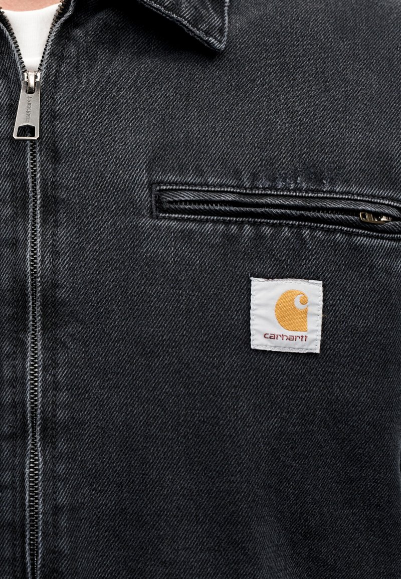 Giacca in denim nera con cerniera argento, tasca sul petto e toppa con il logo Carhartt bianco con un design arancione sul lato sinistro.