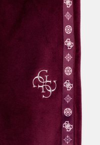 Samtstoff in Mahagoni mit einem zentralen gestickten Logo und einem seitlichen Streifen mit sich wiederholenden grafischen Mustern in Hellrosa.