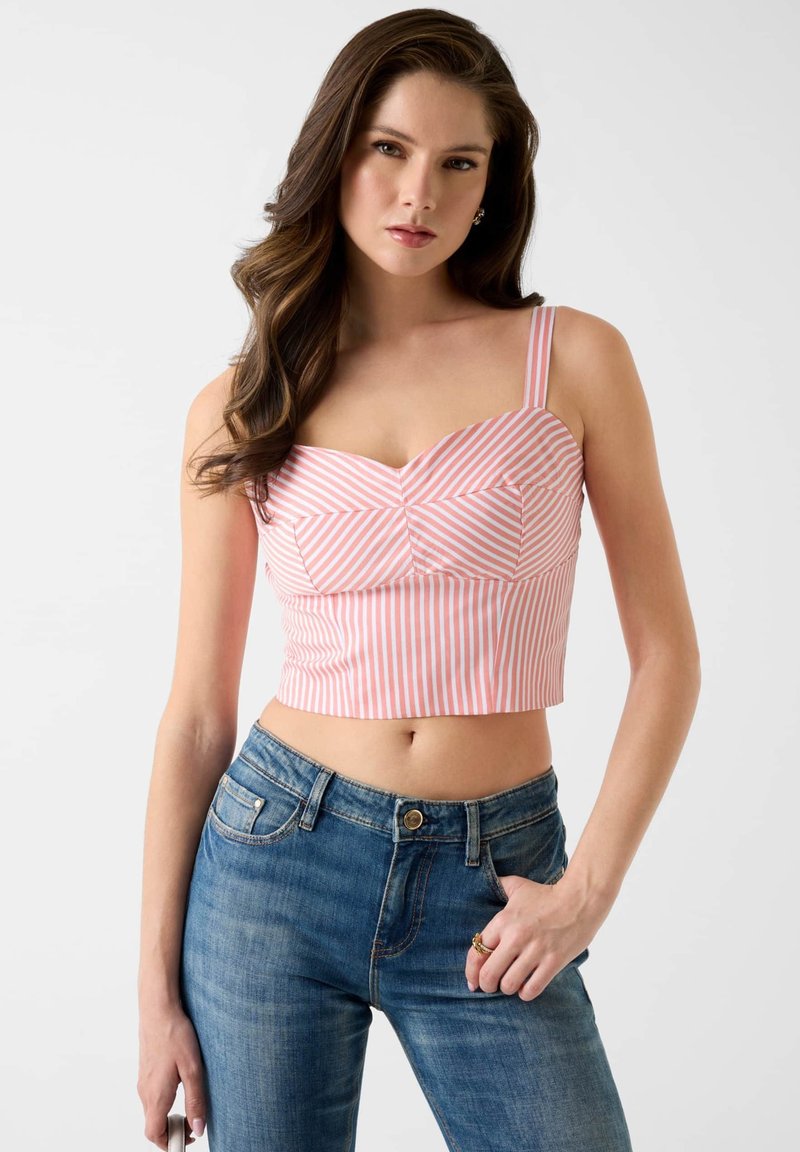 Jeune femme aux longs cheveux bruns portant un crop top rayé rose et blanc et un jean bleu, posant devant un fond blanc.