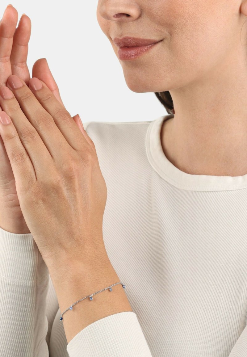 Pulsera de plata con pequeñas piedras preciosas azules, que presenta un diseño de cadena delicado. Lleva puesta sobre un suéter de tonos neutros. Las manos están posicionadas juntas.